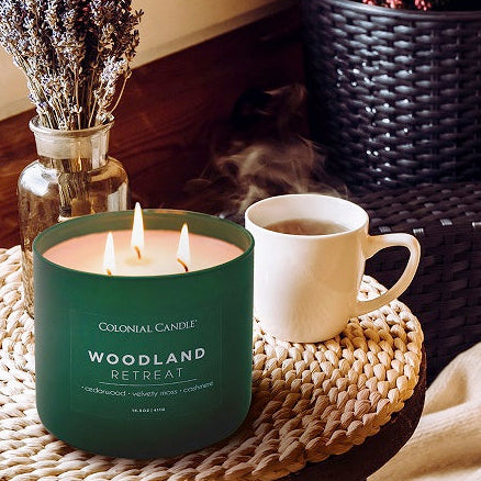 Candle with 'Woodland Retreat' label on a woven table with plants and a cup in a cozy indoor setting.