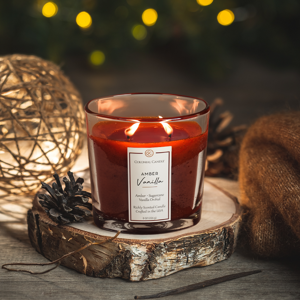 Candle labeled 'Amber Vanilla' on a wooden coaster with decorative lights and pinecones in the background.