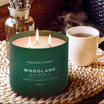 Candle with 'Woodland Retreat' label on a woven table with plants and a cup in a cozy indoor setting.