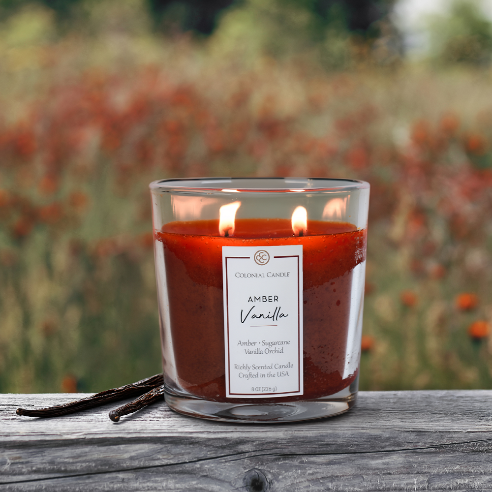 Candle labeled 'Amber Vanilla' with vanilla beans on a wooden surface, blurred natural background