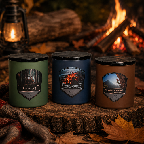 Three scented candles in matte green, blue, and brown jars with black lids are arranged on a rustic wood slice in front of a campfire. Each jar has a shield-shaped label with a forest/fire scene and the text 'M|F'.